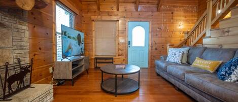 A log cabin interior with a wooden floor, a sofa, a round coffee table, a television, and a stone fireplace.