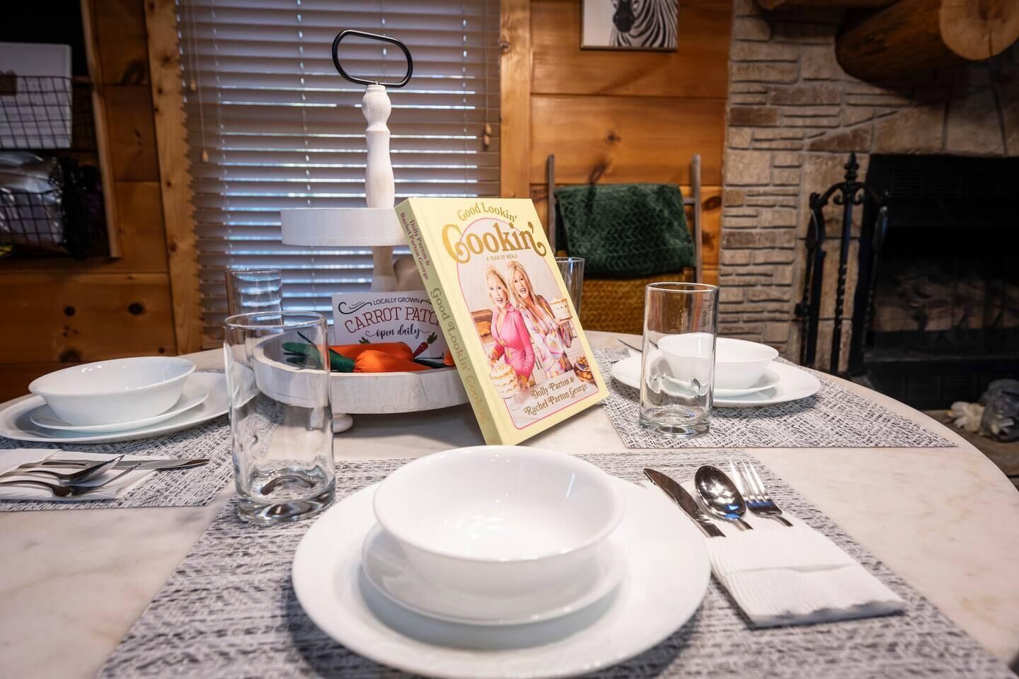 A dining table set with white plates, bowls, and cutlery, a book titled 'Good Lookin' Cookin',' and a bowl of carrots.