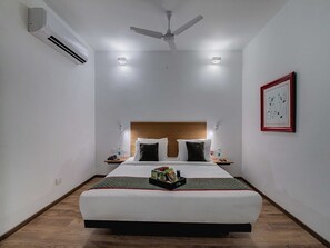 Une chambre d’hôtel avec un lit, un ventilateur de plafond, un climatiseur, des lampes de chevet, un tableau encadré au mur et un plateau avec des collations sur le lit.