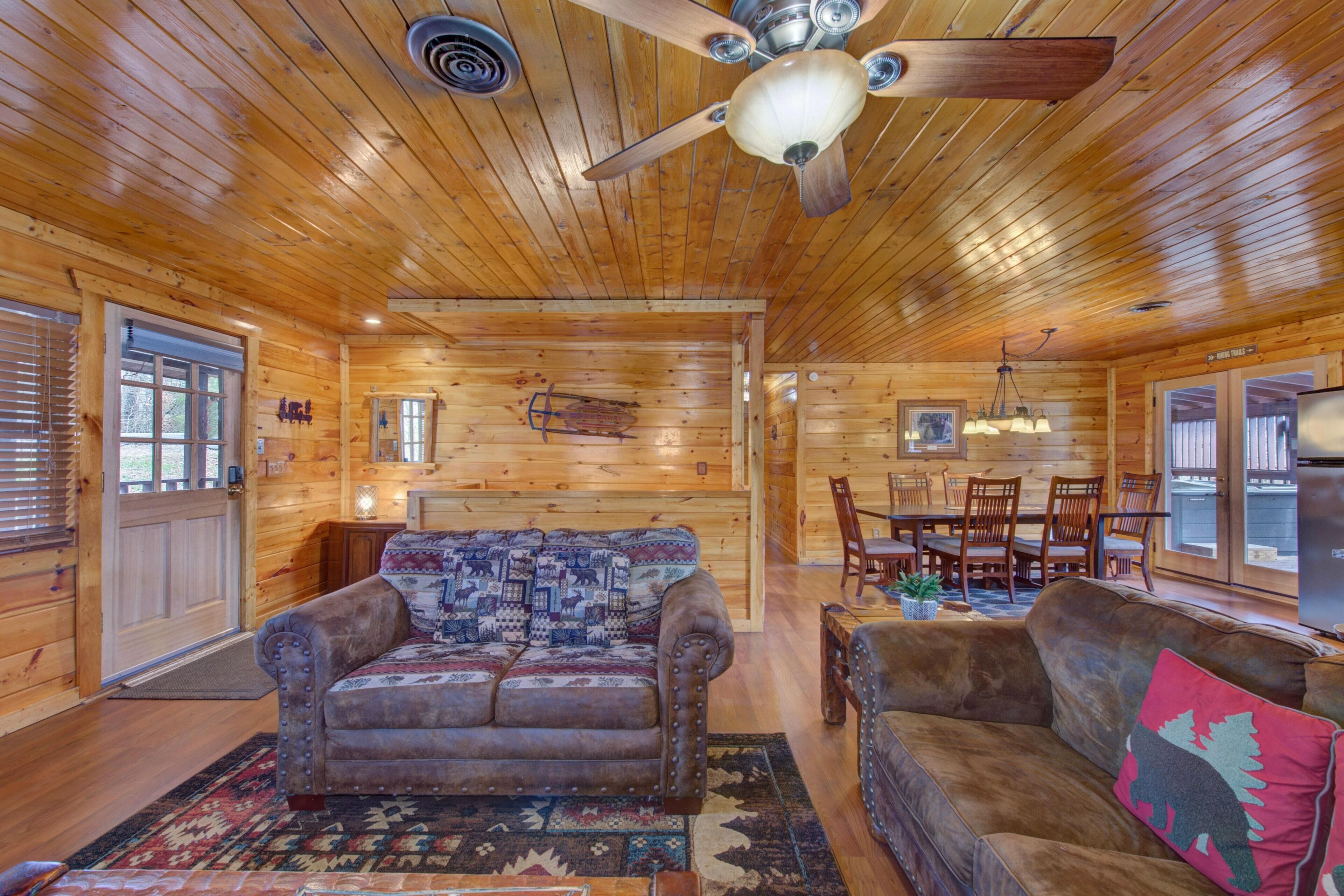 A wooden cabin interior with a ceiling fan, a patterned sofa, a dining area with wooden chairs, and a refrigerator.