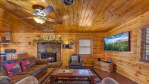 A log cabin interior with a fireplace, a television, a ceiling fan, and a cozy seating area. - Country Cabin in the City (Pigeon Forge)