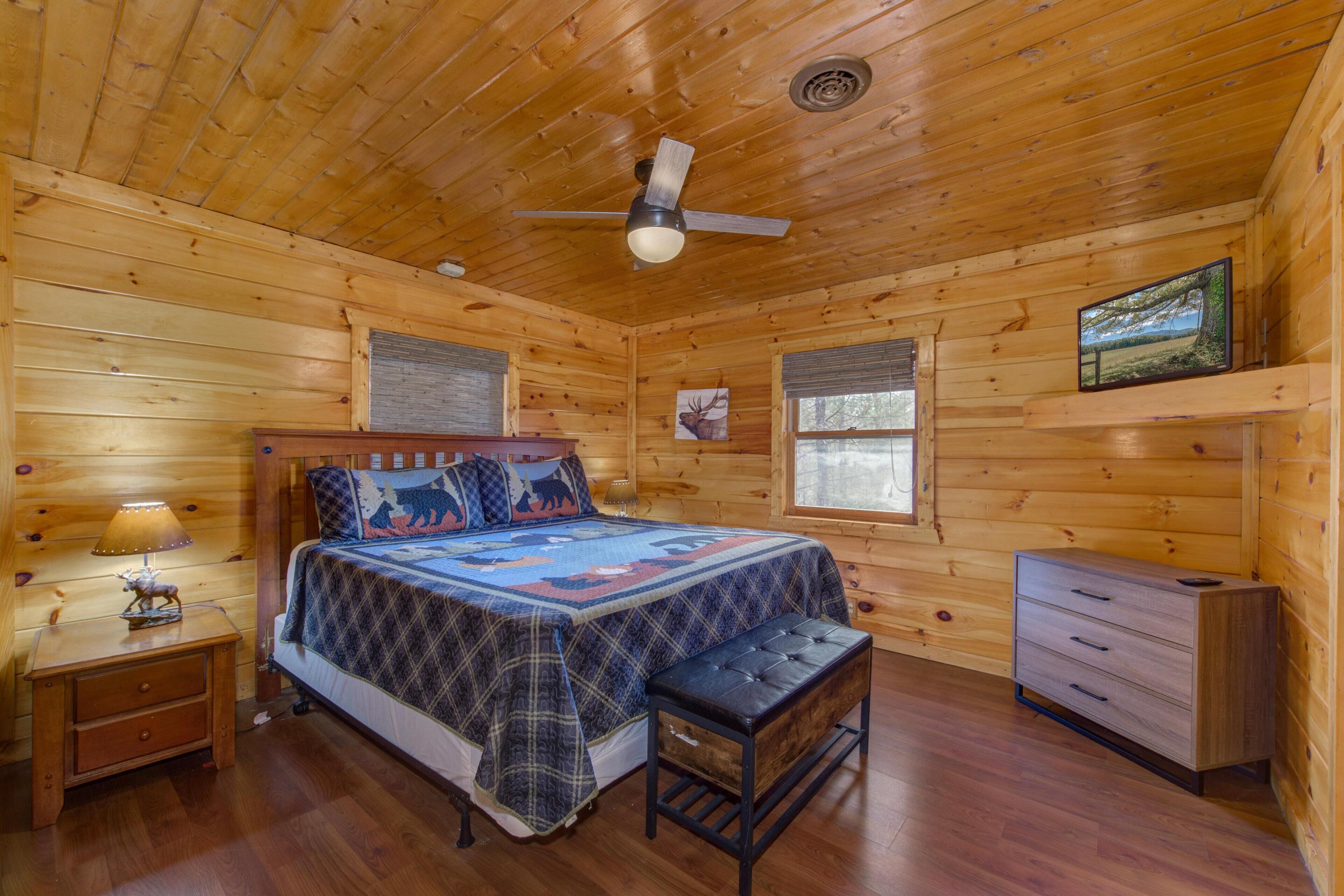 Une chambre à coucher dans une cabane en bois, comprenant un lit, une table de chevet, un banc, une commode et un ventilateur de plafond.