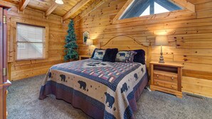 Um quarto de cabana de tronco com uma cama, duas mesas de cabeceira, uma janela com persianas, um pinheiro de Natal e um teto inclinado com claraboia.