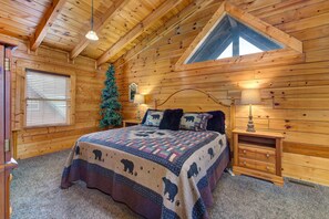 Ein Schlafzimmer in einer Blockhütte mit einem Bett, zwei Nachttischen, einem Fenster mit Jalousien, einem Weihnachtsbaum und einer schrägen Decke mit einem Oberlicht.