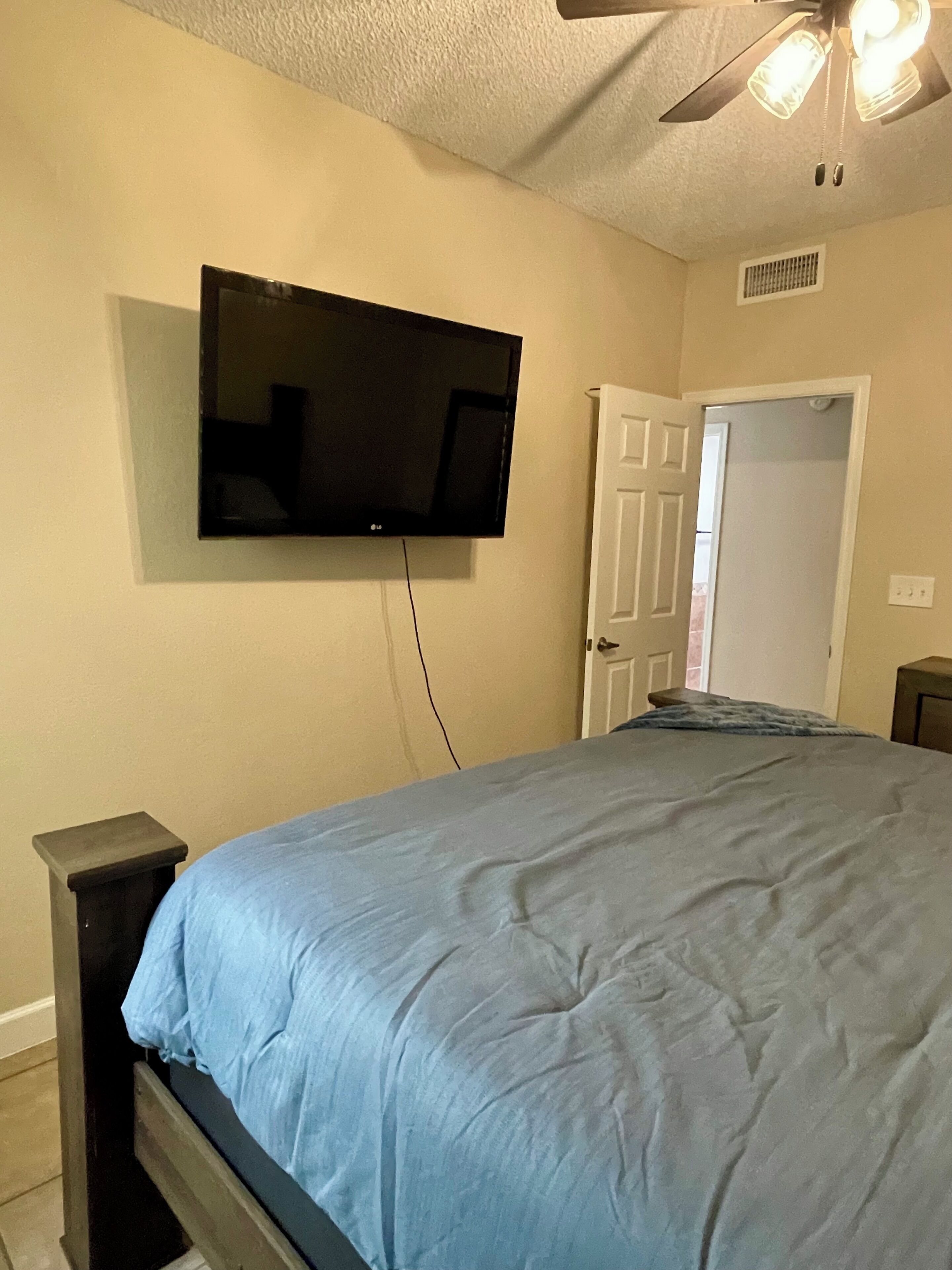 Un dormitorio con una cama, televisión montada en la pared, ventilador de techo y una puerta.