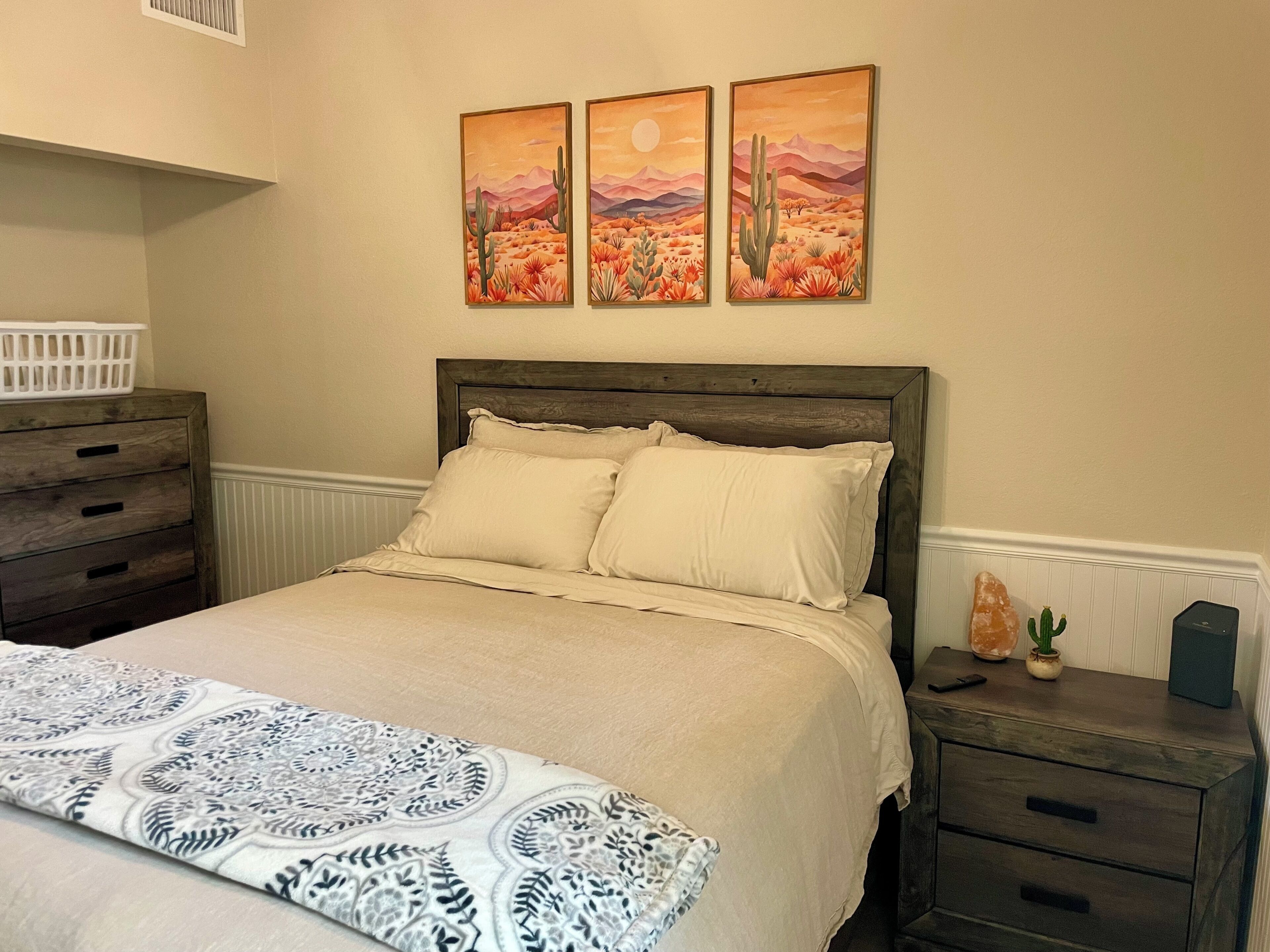 Een slaapkamer met een bed, nachtkastje, kapstok en drie schilderijen aan de muur.