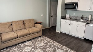 A living room with a beige sofa, a patterned rug, a kitchen with white cabinets, a microwave, and a dishwasher.