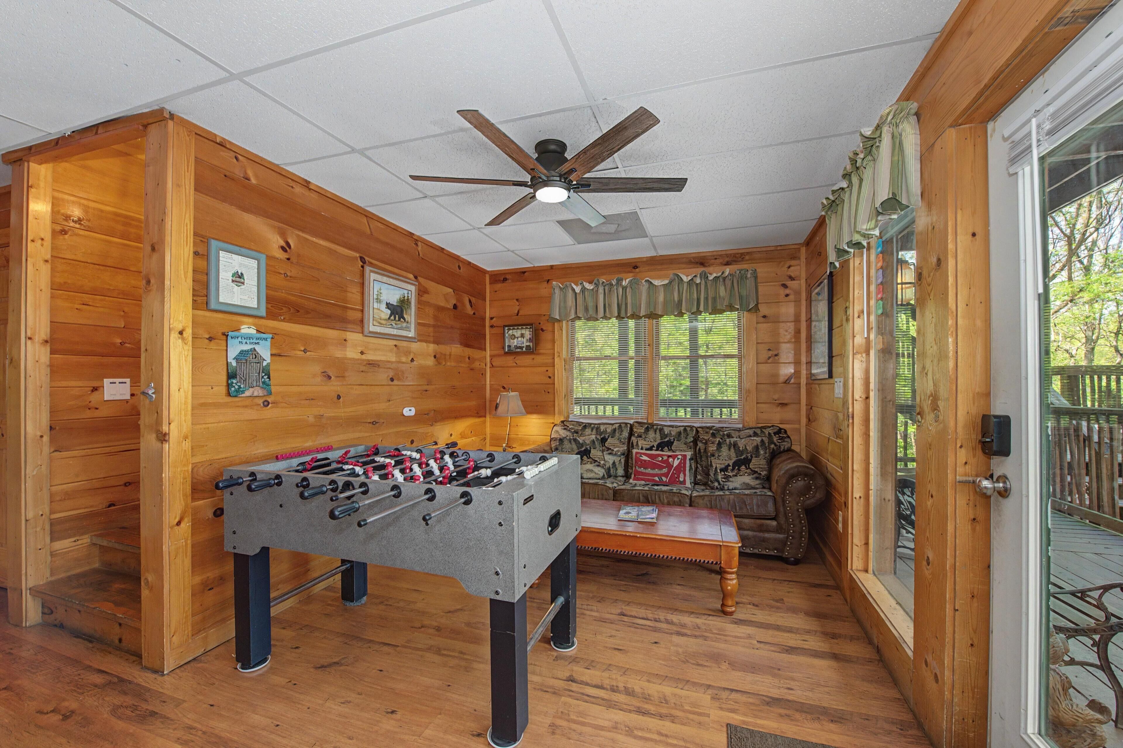 A room with a foosball table, a sofa, a ceiling fan, and a window with a view of greenery.