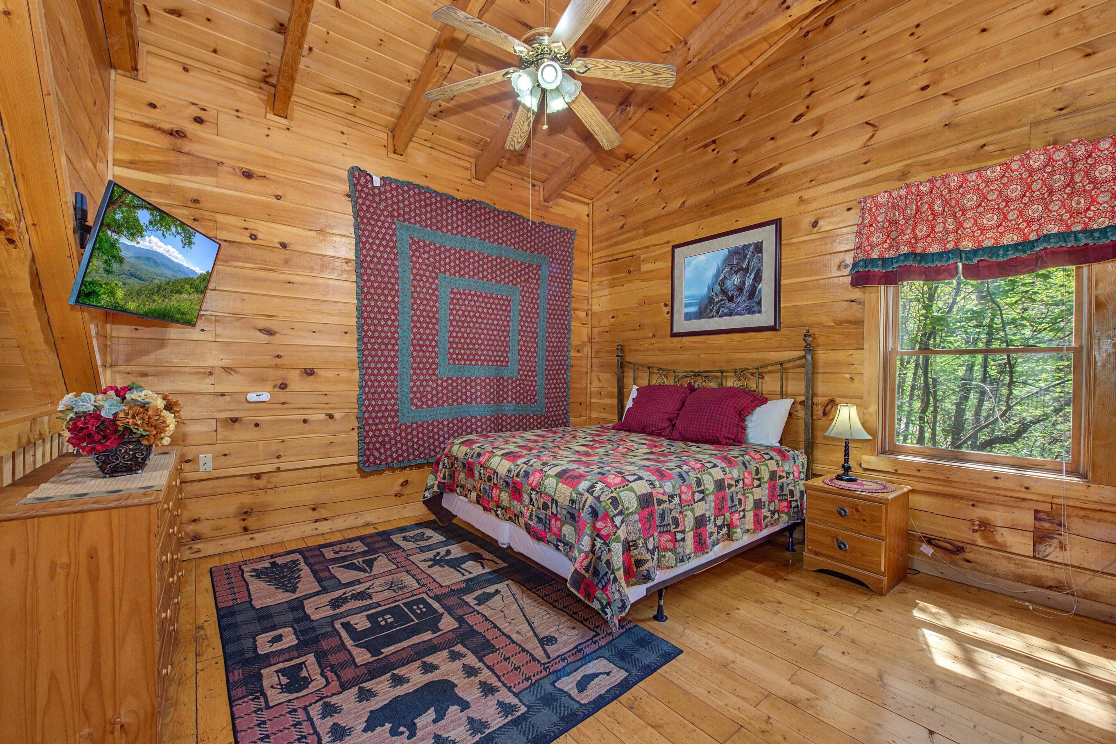 A wooden cabin bedroom with a bed, a television, a window with a view of trees, a ceiling fan, and a quilt on the wall.