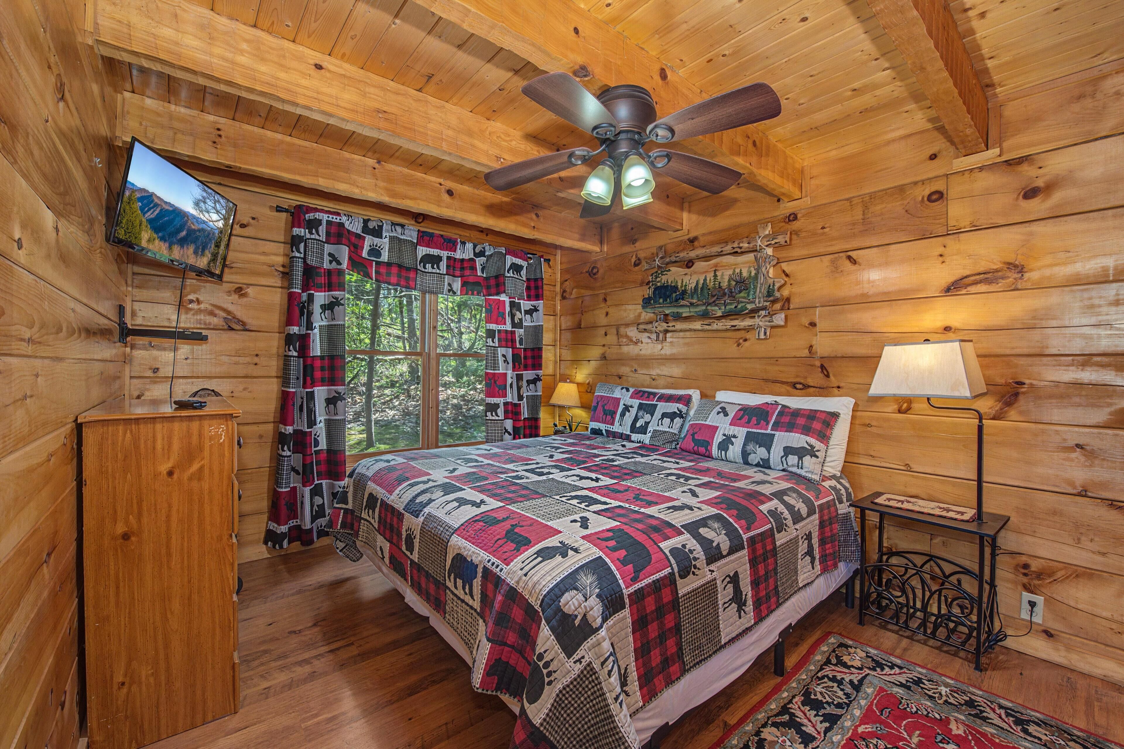 A wooden cabin bedroom with a bed, a ceiling fan, a TV, a dresser, a lamp, and a window with curtains.
