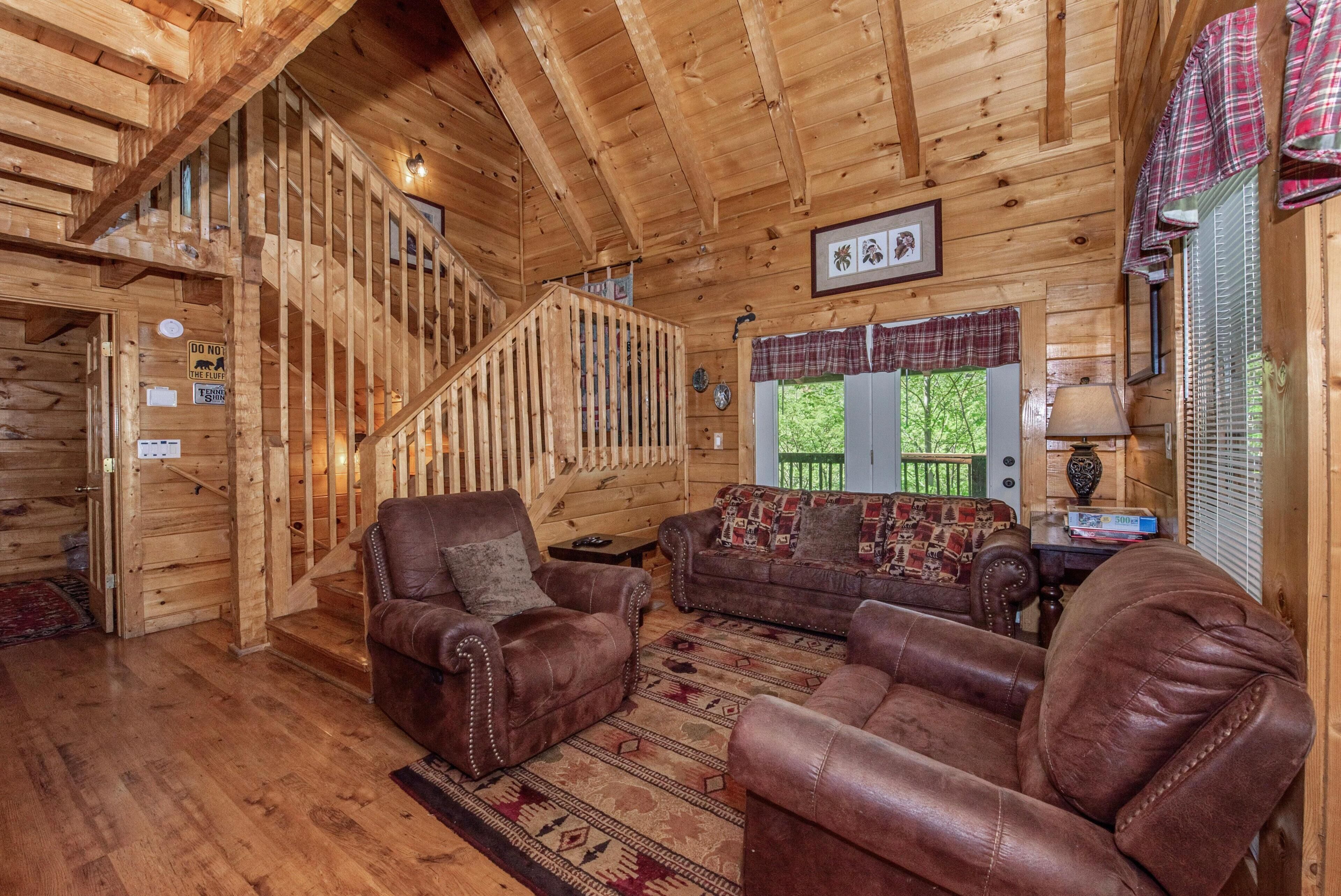 A wooden cabin interior with a staircase, leather sofas, a patterned rug, and a window with blinds.