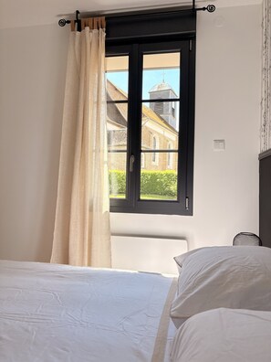A bedroom with a bed, a window with curtains, a chair, and a view of a building outside.