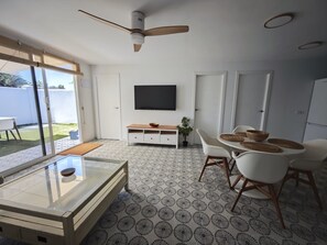 A modern living room with a glass coffee table, white chairs, a wooden TV stand, and a ceiling fan.
