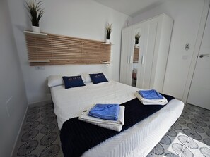 Un lit bien fait, avec des oreillers, une table de chevet sur laquelle sont pliées des serviettes, une armoire et une étagÚre murale agrémentée de plantes.