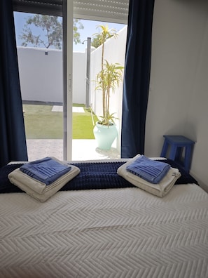 Un lit recouvert dâune couette blanche, avec deux serviettes pliĂ©es, une plante en pot, et une vue sur un mur blanc et un arbre Ă lâextĂ©rieur.