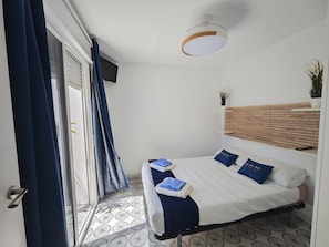 A bedroom with a bed, a ceiling light, a window with curtains, a wall-mounted shelf with a plant, and a patterned floor.