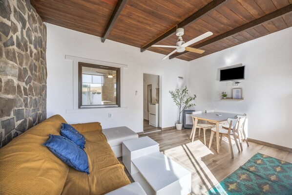 Una sala moderna con pared de piedra, vigas de techo de madera, un sofá con almohadas azules, un comedor con mesa y sillas, y un televisor montado en la pared.