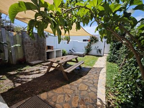 Un patio con un sendero de piedra, una mesa de madera y una zona con sombra gracias a un toldo.