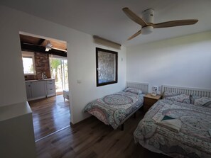 Un dormitorio con dos camas, un ventilador de techo, una obra de arte de piedra en la pared y una cocina visible a través de una puerta abierta.