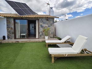 Een klein stenen huis met een zonnepaneel, een schuifdeur naar een keuken en twee rieten loungestoelen op een groen gazon.