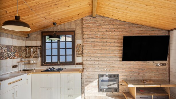 A kitchen with a brick wall, wooden ceiling, and a TV mounted above the counter.