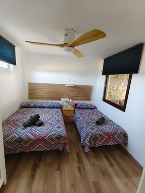 A room with two beds, a ceiling fan, a window with a stone wall view, and a wooden floor.