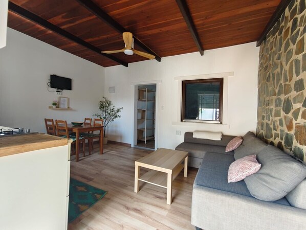 A living room with a wooden ceiling, a stone wall, a sofa with cushions, a wooden table, and a TV mounted on the wall.