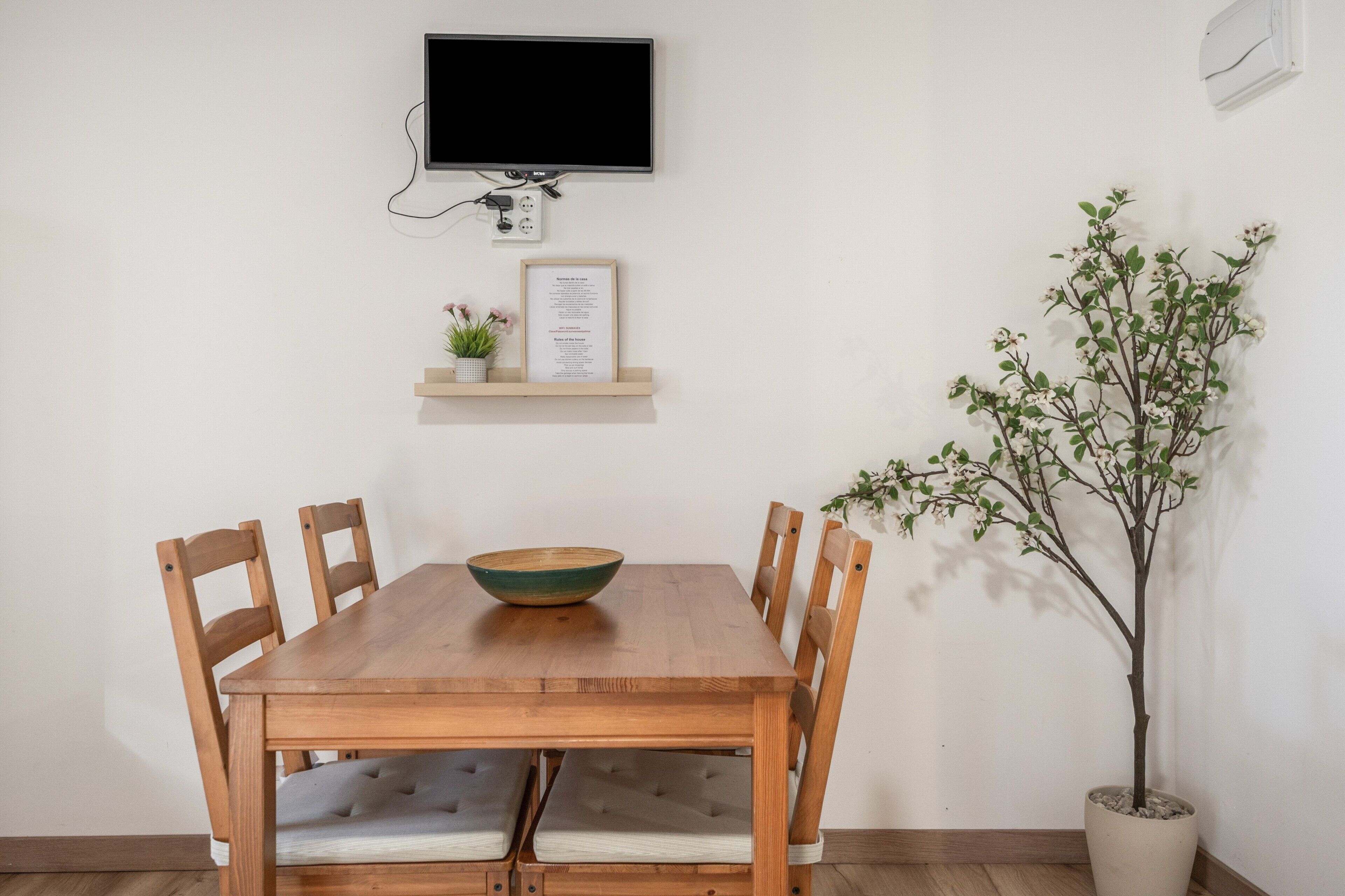 Una mesa de comedor con sillas, un televisor montado en la pared, un estante pequeño con un menú enmarcado y una planta en maceta.