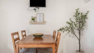 Una mesa de comedor con sillas, un televisor montado en la pared, un estante pequeño con un menú enmarcado y una planta en maceta.