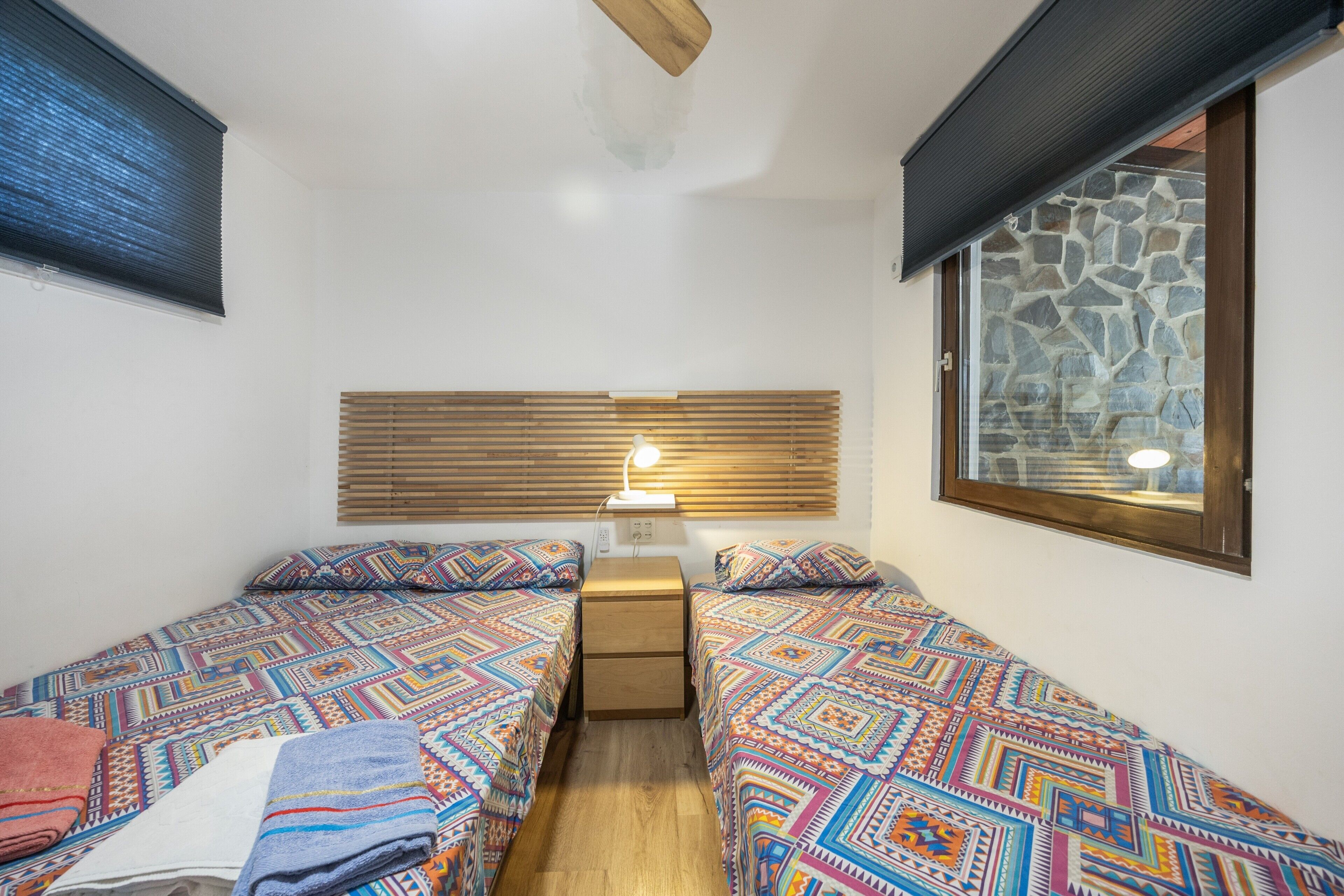 Una habitación pequeña y acogedora con dos camas, una mesita de noche, una lámpara y ventana con vistas a una pared de piedra.