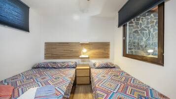 Una habitación pequeña y acogedora con dos camas, una mesita de noche, una lámpara y ventana con vistas a una pared de piedra.
