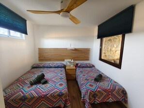 A small, cozy room with two beds, a ceiling fan, a window with a stone wall view, and a bedside table with a lamp.