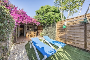 Ein Garten mit einem gepflasterten Weg, zwei Liegestühlen, einem Holzzaun und einer kleinen Terrasse mit Tisch und Stühlen.