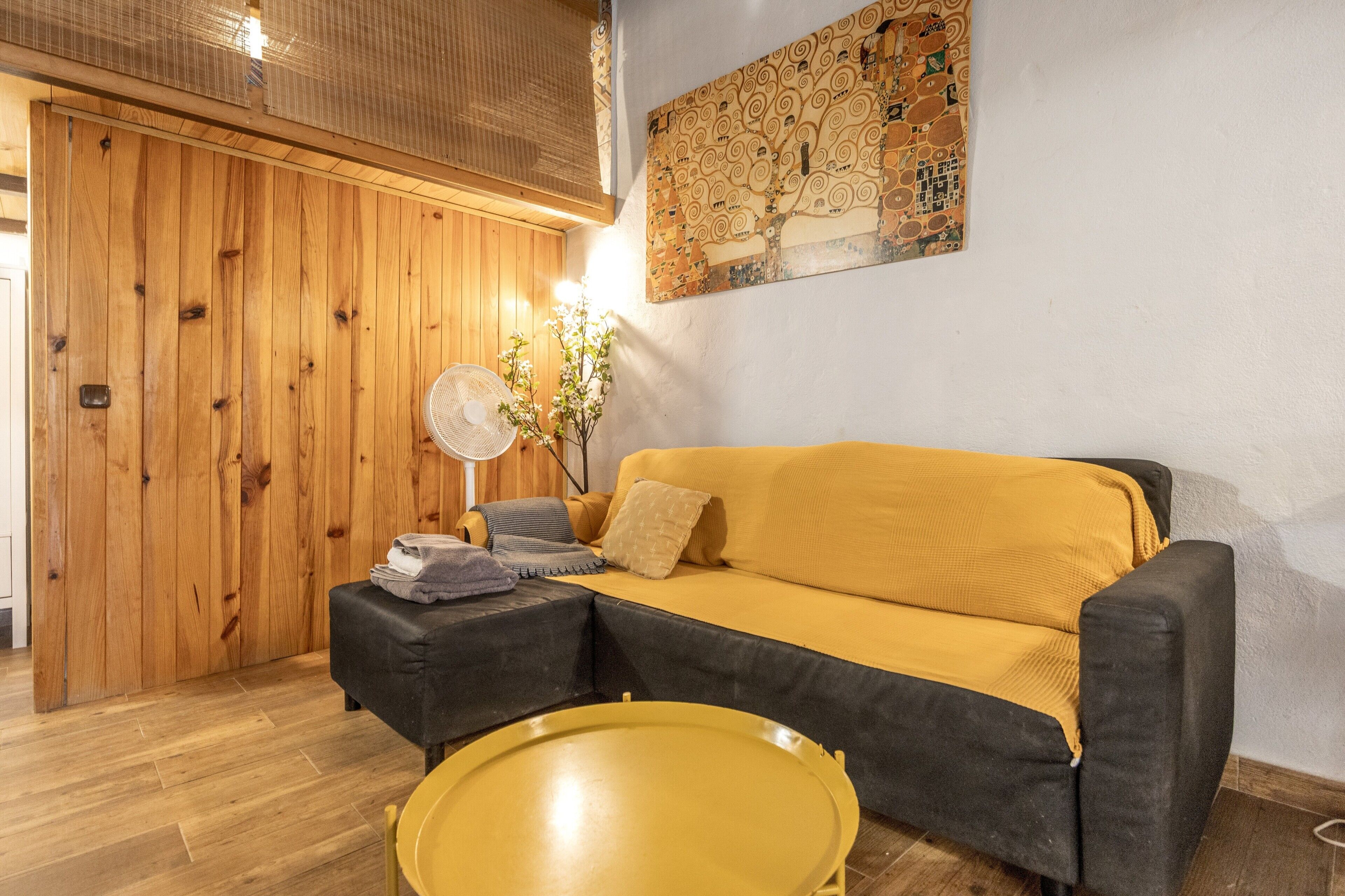 Una habitación con un separador de madera, un sofá amarillo, una mesita y un ventilador.