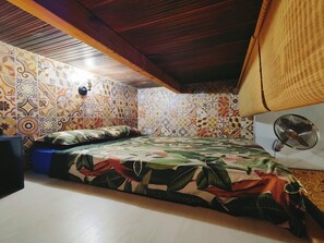 A room with a patterned tile wall, a bed with a tropical print bedspread, a blue trash bin, and a wall-mounted fan.