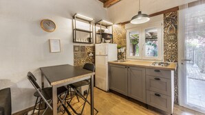 A kitchen with a dining table, chairs, a refrigerator, a shelf with dishes, and a window with a view of greenery. - Casa Pequeña Lulú - Sunwaves, just 5 minutes’ walk from the beach (El Palmar de Vejer)