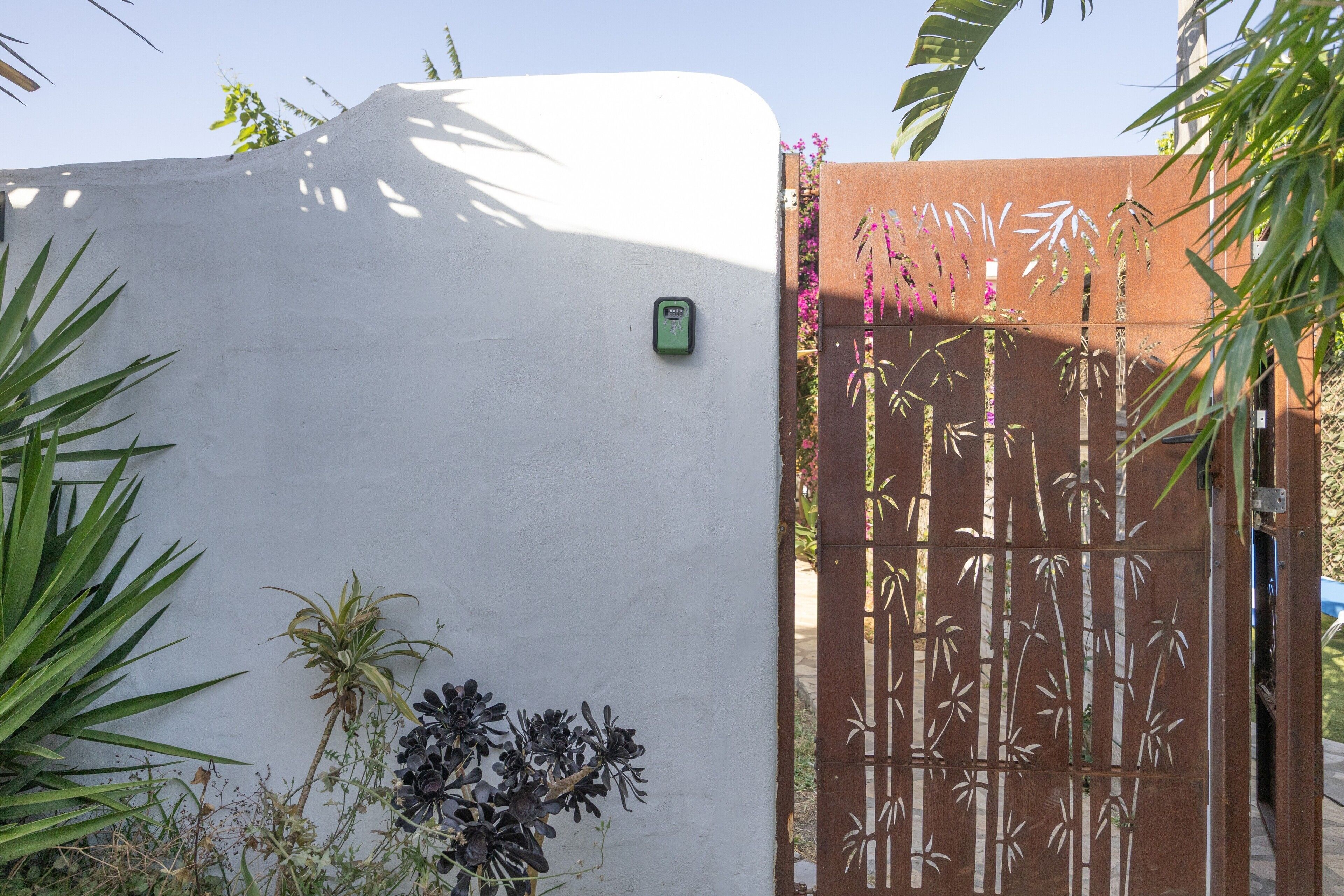 Pared blanca con un buzón verde, un portón con diseño de bambú y diversas plantas.