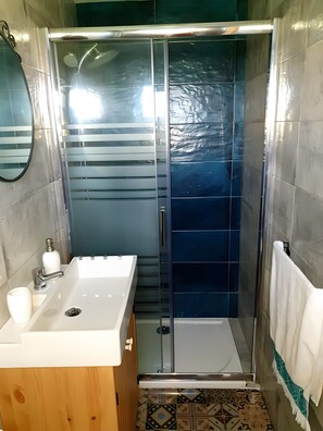 Casa de banho com lavatório branco, espelho, chuveiro com porta de vidro e chão estampado.