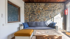 A room with a stone wall, a sofa with cushions, a coffee table, and a window with blinds. - Casa Marina - Sunwaves El Palmar, 5 min from the beach, private garden & Wi-Fi (El Palmar de Vejer)