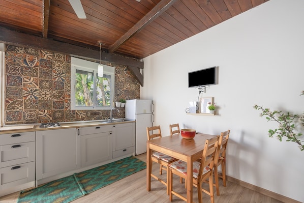 Une cuisine avec des carreaux décoratifs, un plafond en bois, une table à manger avec des chaises, un téléviseur fixé au mur et une petite étagère avec une plante.