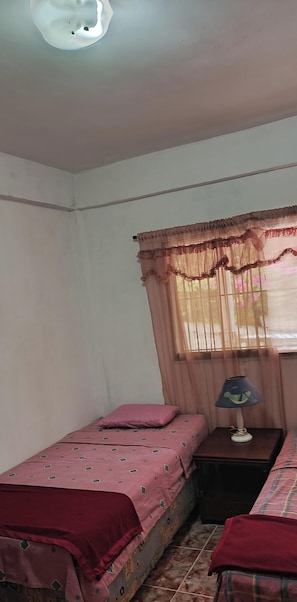 A bedroom with a single bed, a nightstand with a lamp, and a window with sheer curtains.