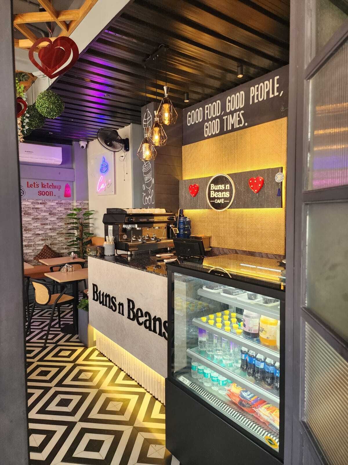 L’intérieur d’un café, avec un comptoir, des machines à café, un présentoir en verre contenant des boissons et un mur orné d’une décoration en forme de cœur et d’un texte.