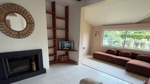 Een woonkamer met een open haard, een televisie op een houten plank, een bank en een raam met uitzicht op groen.