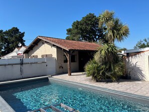 A swimming pool with a tiled deck, a covered outdoor seating area, and a palm tree. - Elegant vacation home - Swimming pool, close to lake, cycle path and shops (Biscarrosse)