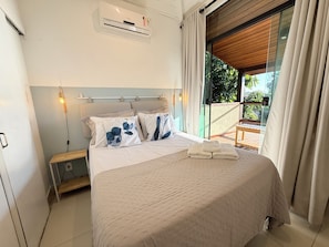 Ein Schlafzimmer mit Bett, Nachttisch, Klimaanlage und Blick auf Grün hinter einer Glastür.