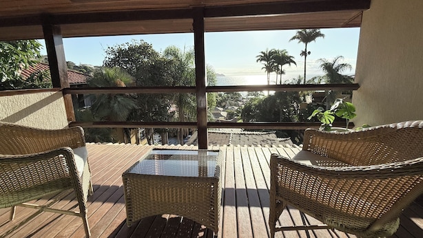 Un terrazzo in legno con mobili in vimini e un tavolo con piano in vetro, con vista su una zona costiera con palme e edifici.