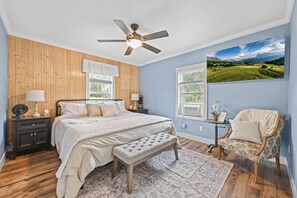 Un dormitorio con cama, un banco, una silla, un ventilador de techo y un ventanal con vistas al paisaje.