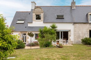 Une maison à deux étages avec une façade en pierre, un toit en tuiles et un petit porche aménagé avec du mobilier d’extérieur.