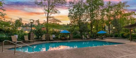 Una piscina con lettini e ombrelloni, immersa nel verde e illuminata dal cielo al tramonto.