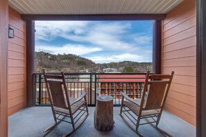 Un balcon avec deux fauteuils à bascule et une petite table en bois, d’où l’on peut admirer un bâtiment aux toits rouges et un paysage vallonné.